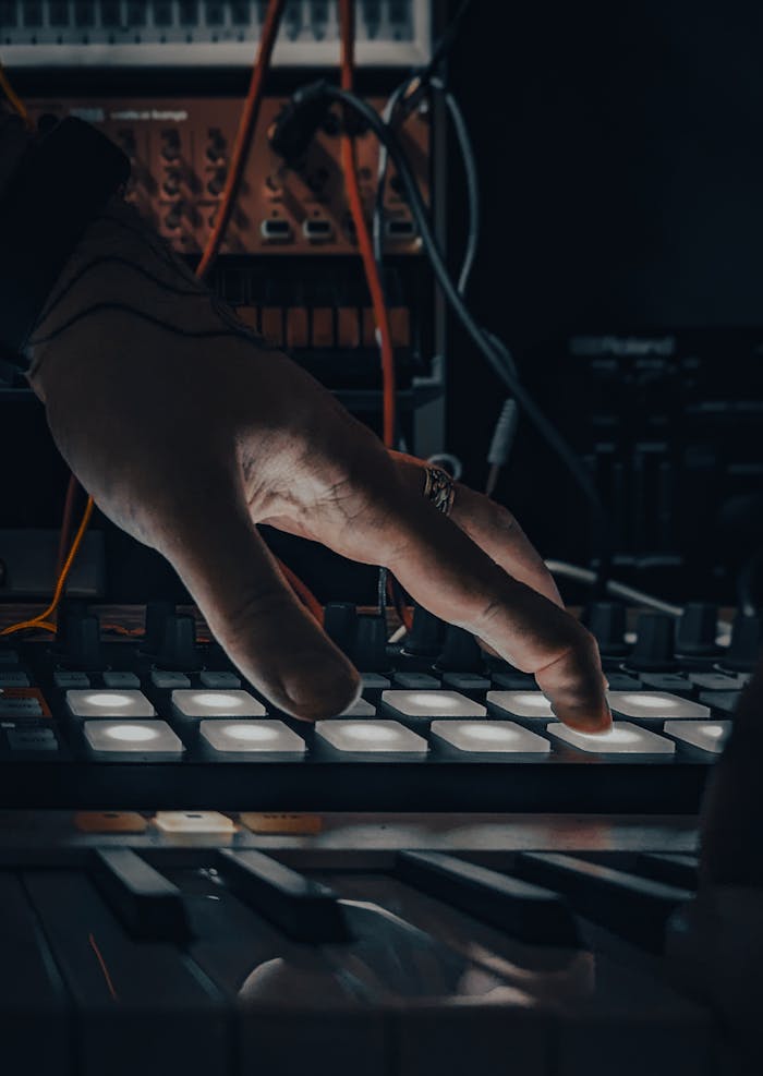 A musicians hand operating a synthesizer pad with glowing buttons in a dark studio.